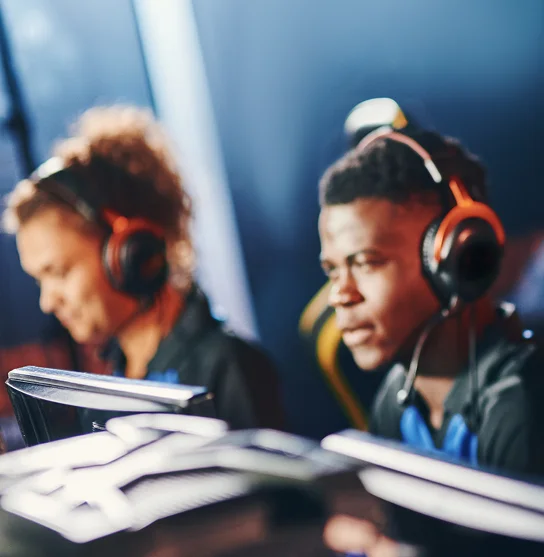 Quatre joueurs assis côte à côte, équipés de casques et vêtus de t-shirts bleus, sont plongés dans leurs écrans dans une pièce faiblement éclairée, faisant preuve d'esprit d'équipe et de concentration